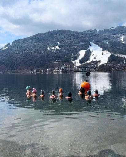 Seven people standing waist-deep in a calm lake with snow-covered mountains in the background under a partly cloudy sky.