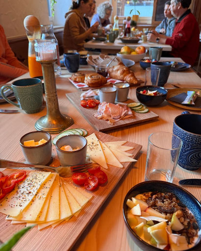 Table set with wooden boards of assorted sliced cheese, cherry tomatoes, cucumber, bread rolls, croissants, and cold cuts, with people conversing in the background.