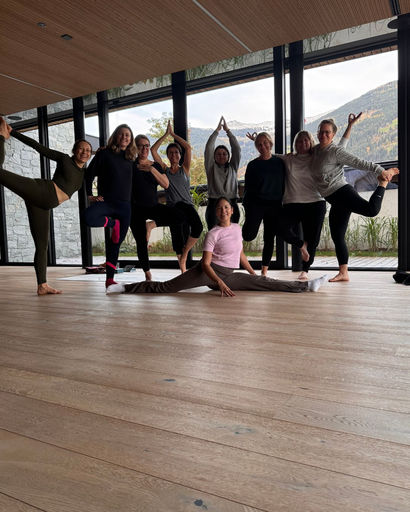 Group of nine women practicing yoga indoors near large windows with mountain view, performing various poses including splits and balancing postures.