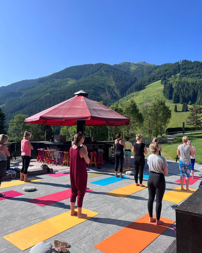 Group of people standing on colorful yoga mats outdoors with a red umbrella and mountains in the background.