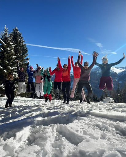 Group of people in winter clothing jumping and celebrating on snow with pine trees and mountains in the background under clear blue sky.