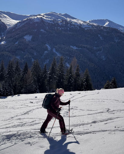 Person snowshoeing on snow-covered terrain with mountains and pine trees in the background under a clear blue sky.