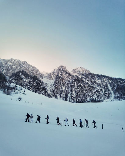 Group of hikers walking in a snowy landscape with snow-covered mountains and a clear sky in the background.