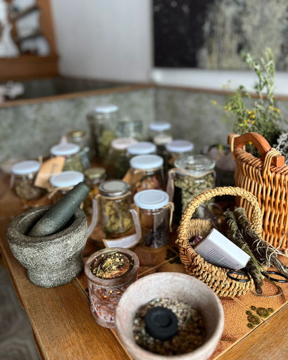 Table with various jars of dried herbs, a stone mortar and pestle, a basket with herbs and a small grinder.