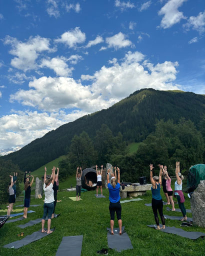 Group of people practicing yoga outdoors on mats on grass with mountains and blue sky in background.
