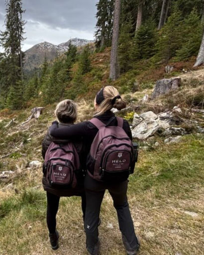 Two hikers with matching backpacks and black outfits, standing arm in arm on a grassy trail with trees and mountains in the background.