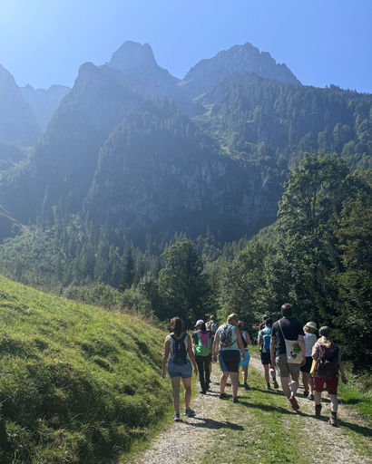 Group of hikers walking on a grassy trail surrounded by trees with tall, rocky mountains in the background under a clear blue sky.