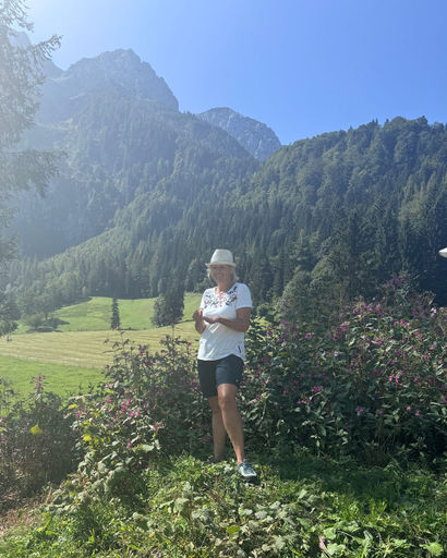 Person wearing a hat and casual clothes standing on green grass with purple flowers in front of forested mountains under a clear blue sky.