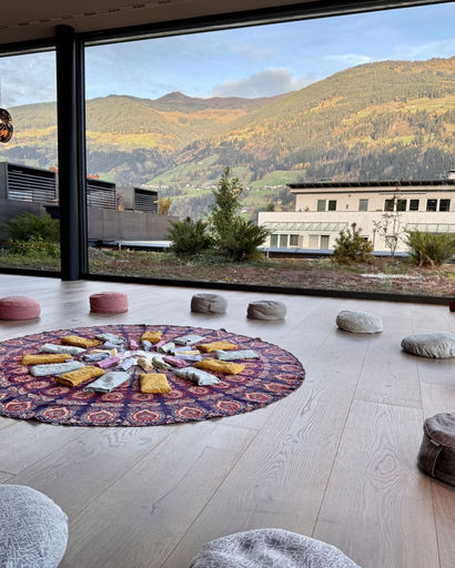 Circular patterned rug with arranged candles and cushions on light wooden floor, large window shows mountainous landscape outside.