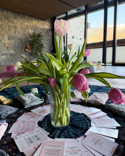 Glass vase with pink tulips placed on a table surrounded by arranged cards in a cozy room with stone wall and large windows.