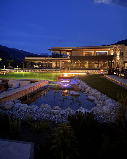 Modern house at dusk with large glass windows, outdoor pool, and a fire pit surrounded by rocks and landscaping.