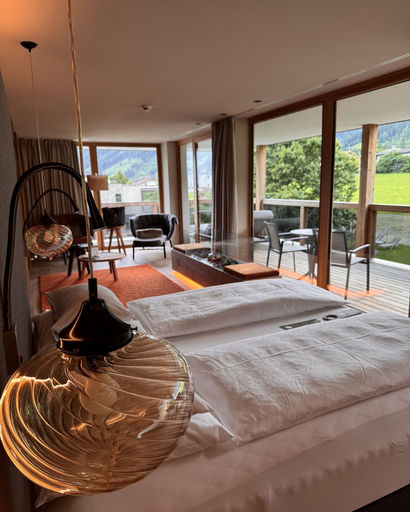 Modern hotel room with two white beds, hanging glass lamps, a seating area, and large windows opening to a balcony with outdoor chairs overlooking greenery.