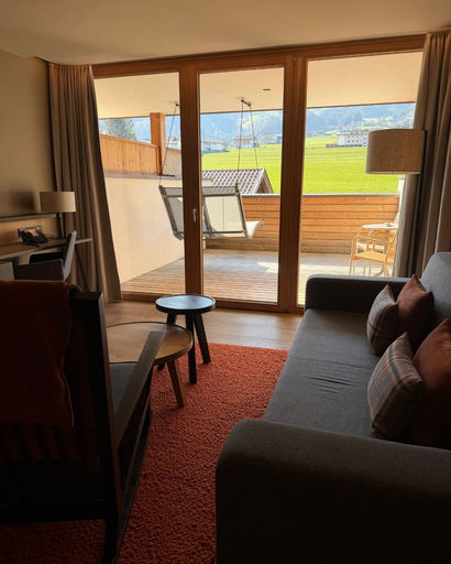 Cozy living room with sofa, pillows, armchair, round tables, and a desk lamp, opening to a balcony with hanging chair and green fields beyond.