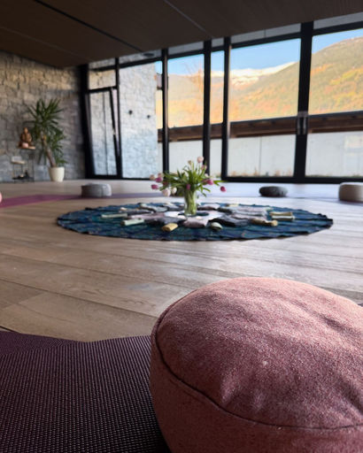 Cozy meditation room with floor cushions arranged around a circular rug centerpiece decorated with flowers, large windows revealing a mountain view, and natural light.