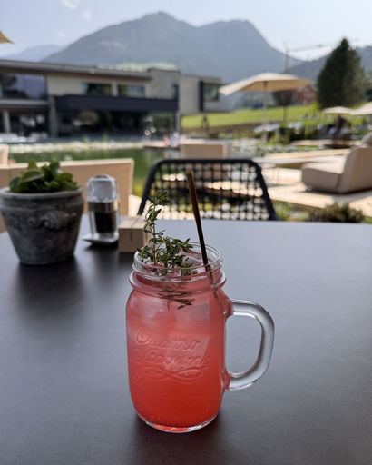 Pink refreshing drink with herb garnish and a straw in a mason jar on a dark table with outdoor seating and mountain scenery in the background.