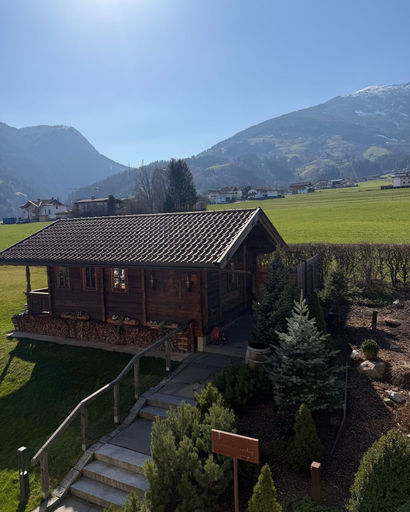 Wooden cabin with stacked firewood under the porch, surrounded by green shrubs and a pathway, set against a backdrop of green fields and distant mountains under a clear blue sky.