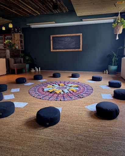 Circular arrangement of black meditation cushions and scattered papers around a colorful patterned cloth on a wooden floor in a room with dark walls and plants.