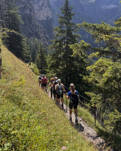 Group of hikers walking in single file along a narrow mountain trail surrounded by green trees and steep cliffs.