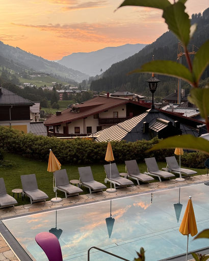 Outdoor pool with yellow umbrellas and lounge chairs at sunset, overlooking a village and forested mountains in the background.
