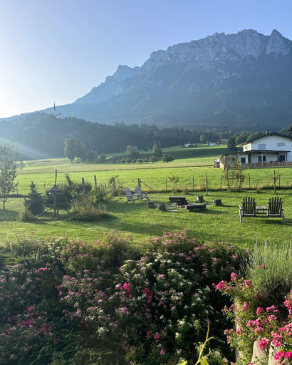 Sunny rural scene with green grass, garden flowers, wooden chairs, and a white house set against a mountain backdrop.