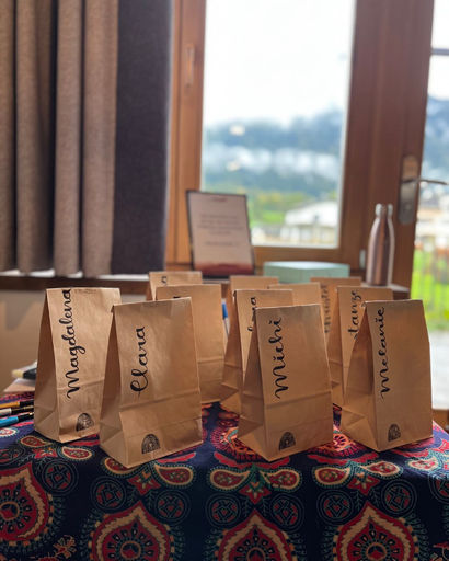 Brown paper bags with handwritten names placed on a colorful patterned tablecloth by a window.