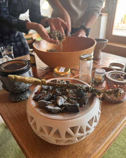 Hands mixing dried herbs over a large wooden bowl on a table with various jars and a smudge stick in a carved pot.