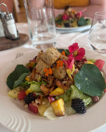 Fresh salad with mixed greens, blackberries, croutons, edible flowers, and diced yellow and red fruits on a white plate.