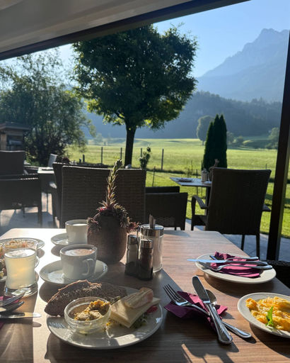 Outdoor table set with breakfast dishes, drinks, and cutlery overlooking a grassy field, trees, and mountains under a clear sky.