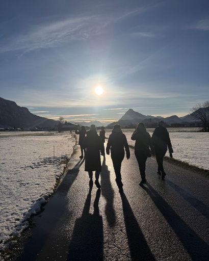 Four people walking on a paved path with snow-covered fields on each side under a bright sun and clear sky, casting long shadows.