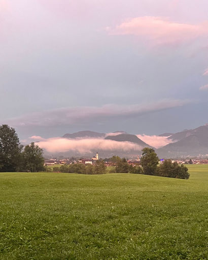 Green field with trees in foreground, a village with a church and mountains partially covered by low clouds in the background under a pinkish sky.
