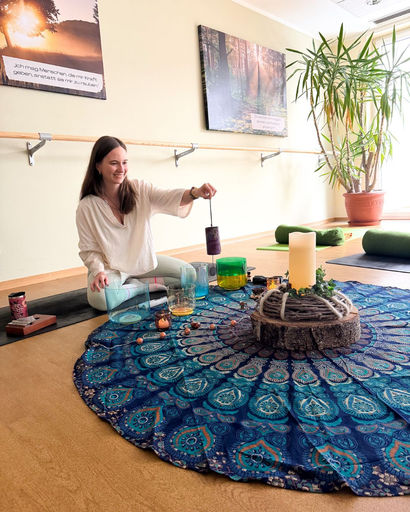 Woman sitting on the floor in a bright room, playing a purple singing bowl over a colorful blue patterned cloth with candles and glass bowls arranged on it.
