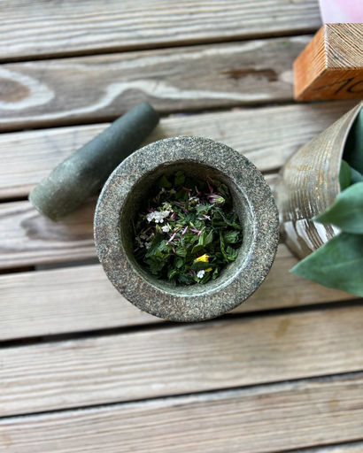 Top view of a stone mortar with fresh herbs and flowers inside and a pestle resting on wooden planks.
