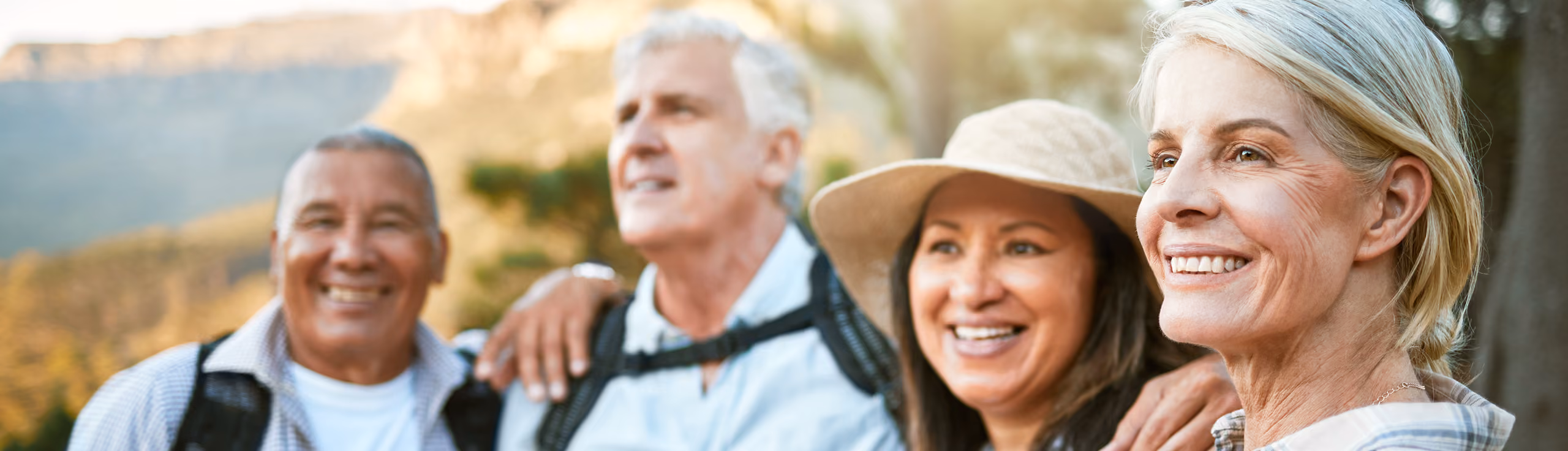 Group of four smiling mature friends outdoors with mountain background, two men and two women, one wearing a hat.