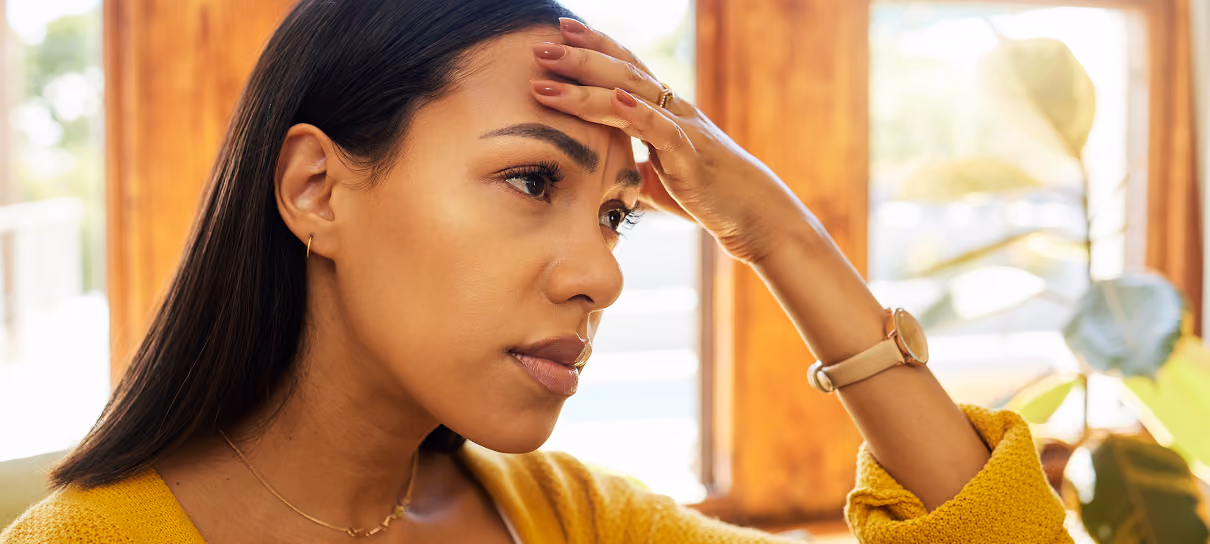 Woman in yellow sweater holding her forehead, looking worried or stressed indoors near a window.