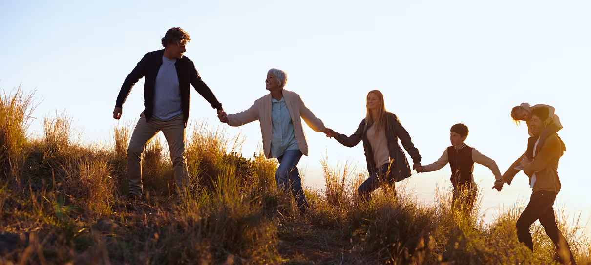 Multigenerational family holding hands and walking up a grassy hill at sunset.