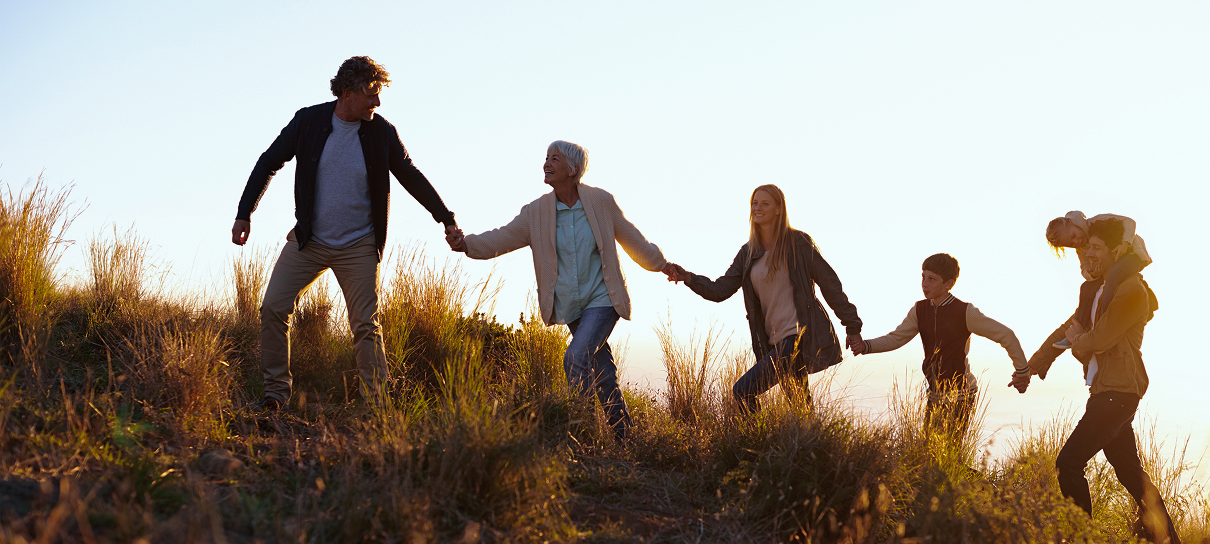 Multigenerational family holding hands and walking up a grassy hill at sunset.
