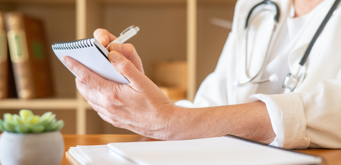 Doctor writing notes on a small spiral notebook with a stethoscope around their neck.