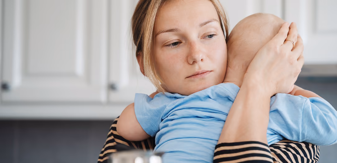 A woman holding and comforting a baby dressed in a blue shirt in a kitchen setting.