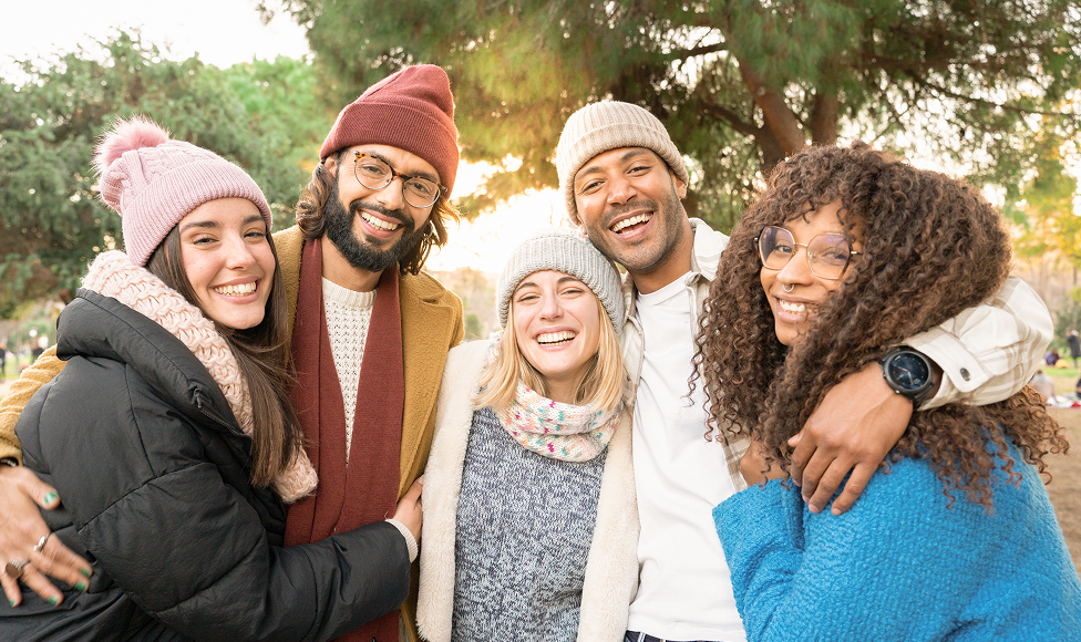 A group of five diverse friends wearing winter clothing, smiling and hugging outdoors with trees in the background.