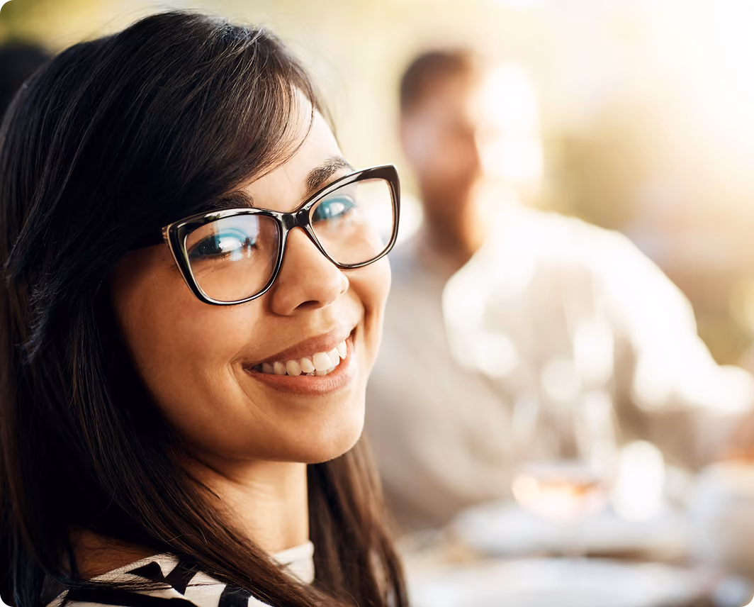 Smiling woman with glasses looking at the camera in a sunlit indoor setting with a blurred man in the background.