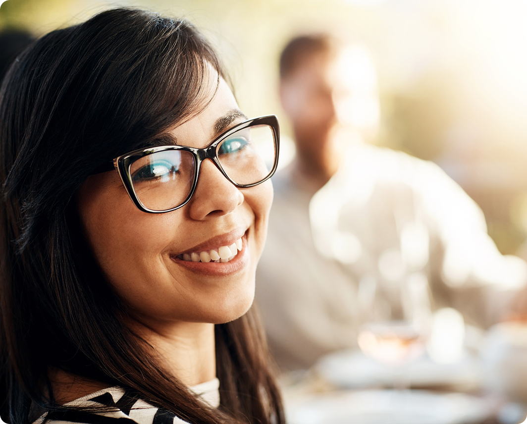 Smiling woman with glasses looking at the camera in a sunlit indoor setting with a blurred man in the background.