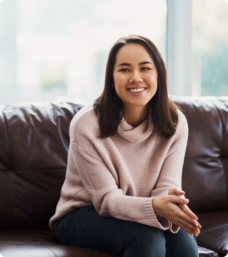 Smiling young woman with dark hair wearing a cozy pink sweater sitting on a brown leather couch.