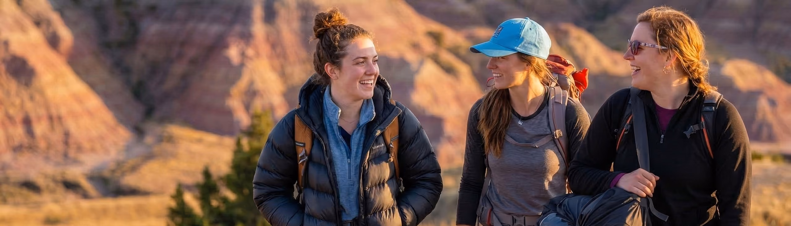 Three women smiling and talking while hiking outdoors with backpacks against a backdrop of sunlit rocky mountains.
