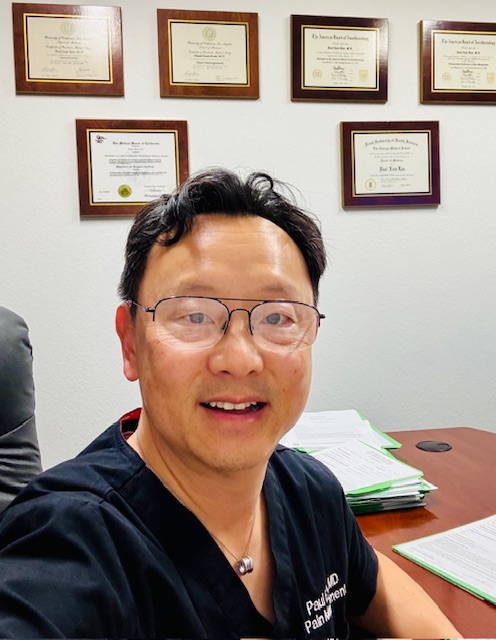Smiling male doctor with glasses sitting at a desk in an office with framed medical certificates on the wall behind him.