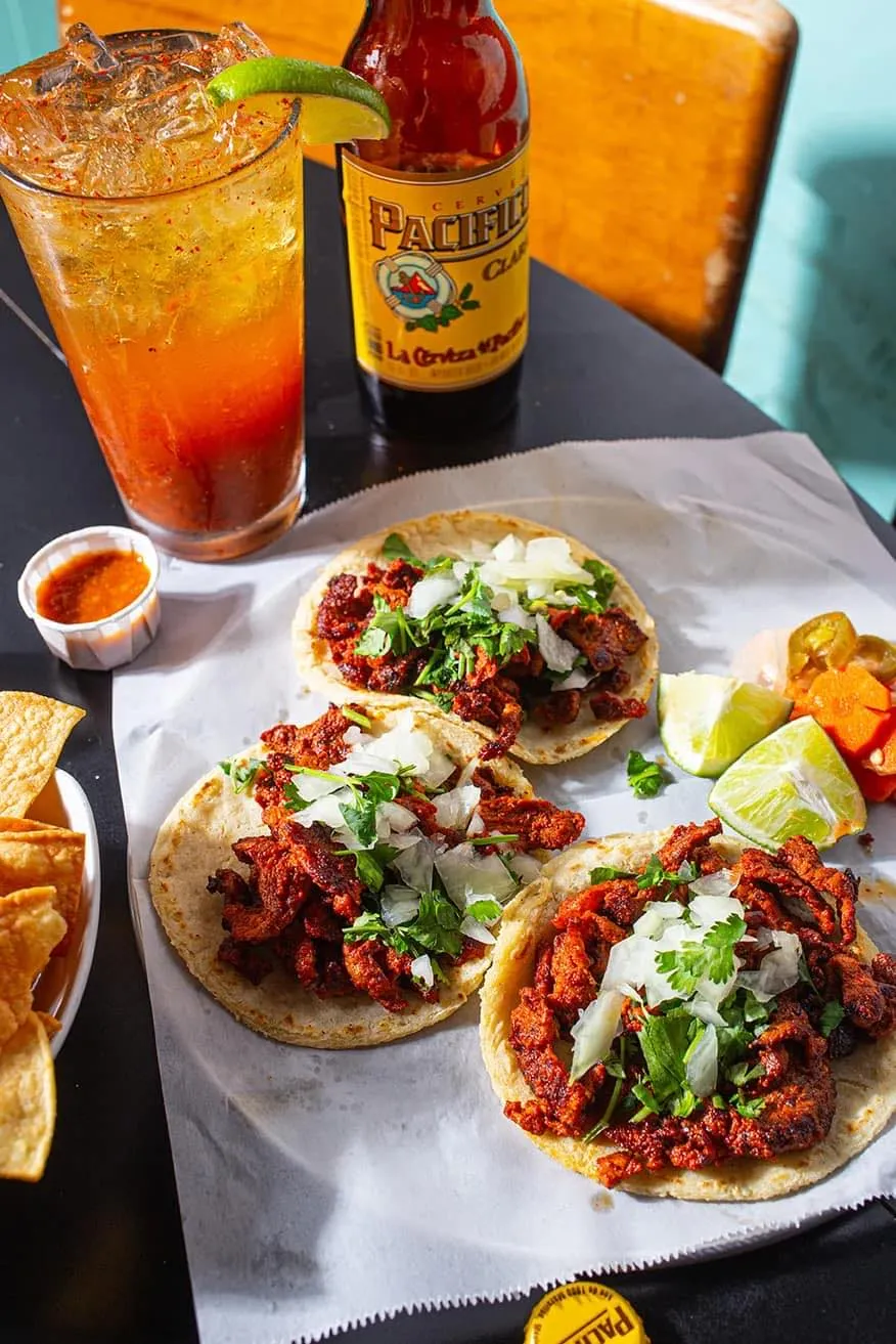 Three tacos with seasoned meat, chopped onions, and cilantro on a white paper with lime wedges, a bottle of Pacifico beer, a michelada cocktail glass garnished with lime and chili powder, and chips with salsa on the side.