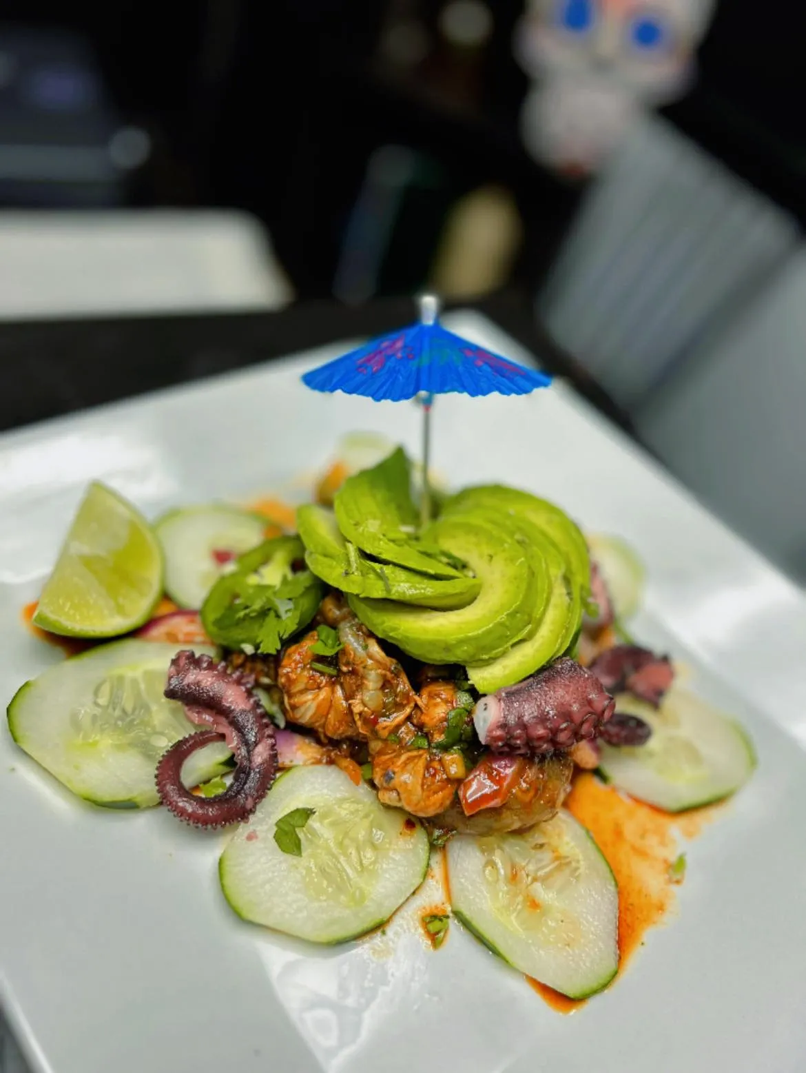 Plated seafood dish with octopus tentacles, sliced avocado, cucumber rounds, a lime wedge, and garnished with a small blue cocktail umbrella.