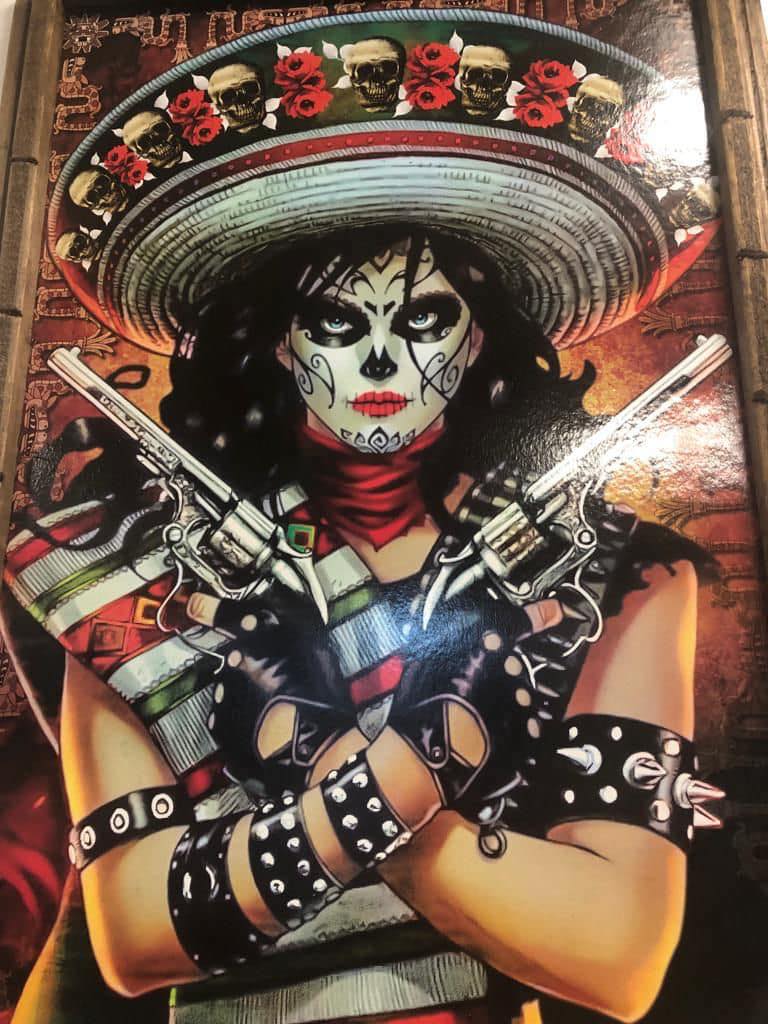 Woman with Day of the Dead face paint wearing a large hat decorated with skulls and roses, crossed arms holding two revolvers, and studded black gloves and armbands.