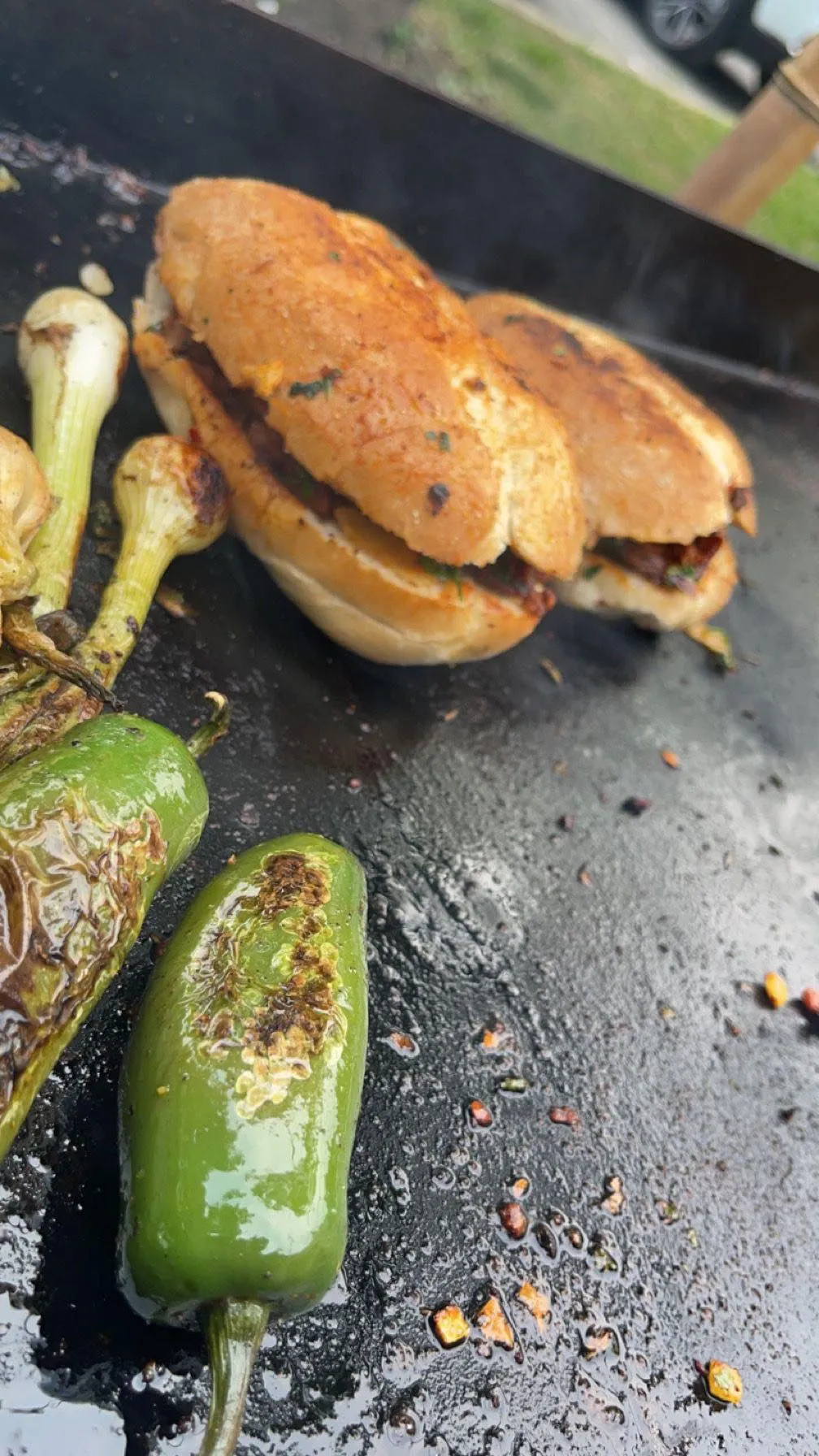 Two toasted sandwiches and charred green peppers cooking on a flat top grill with grilled spring onions.
