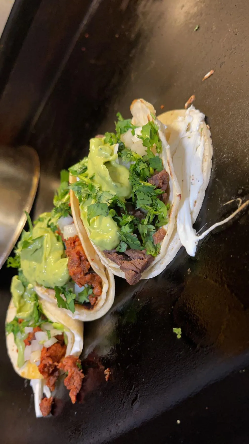 Three tacos on a griddle topped with chopped onions, cilantro, guacamole, and creamy white sauce, with different meat fillings.
