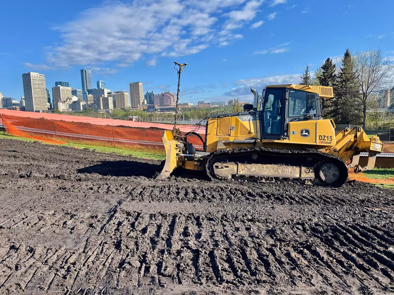 Yellow bulldozer on a muddy construction site with a city skyline and trees in the background under a blue sky.